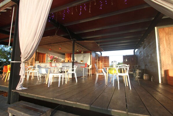 Un espacio exterior para comer con mesas y sillas de madera, una zona de bar con taburetes, y vista al interior a través de un ventanal grande.