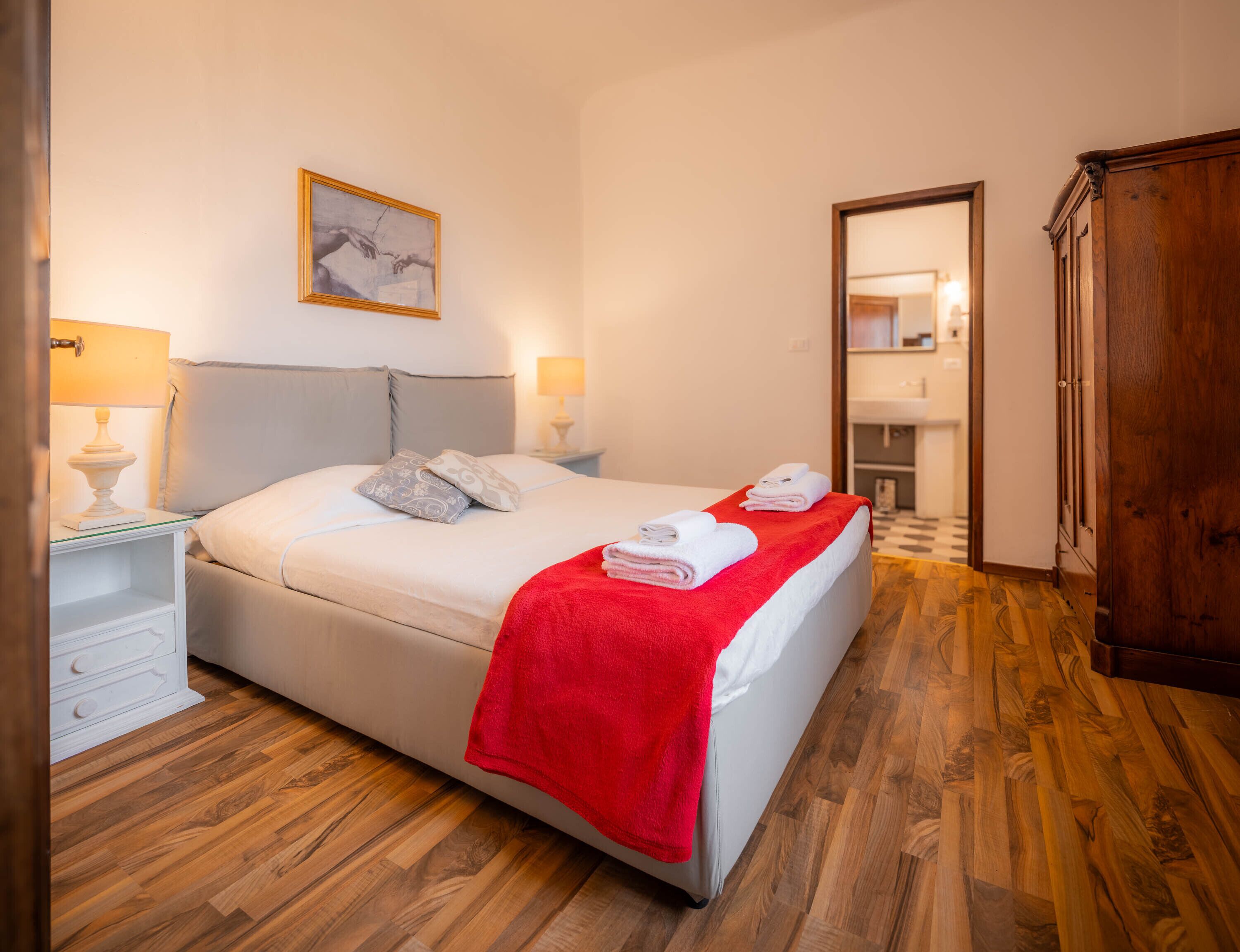 Un lit bien fait, recouvert d’une couverture rouge, une table de chevet avec une lampe et un tableau accroché au mur.