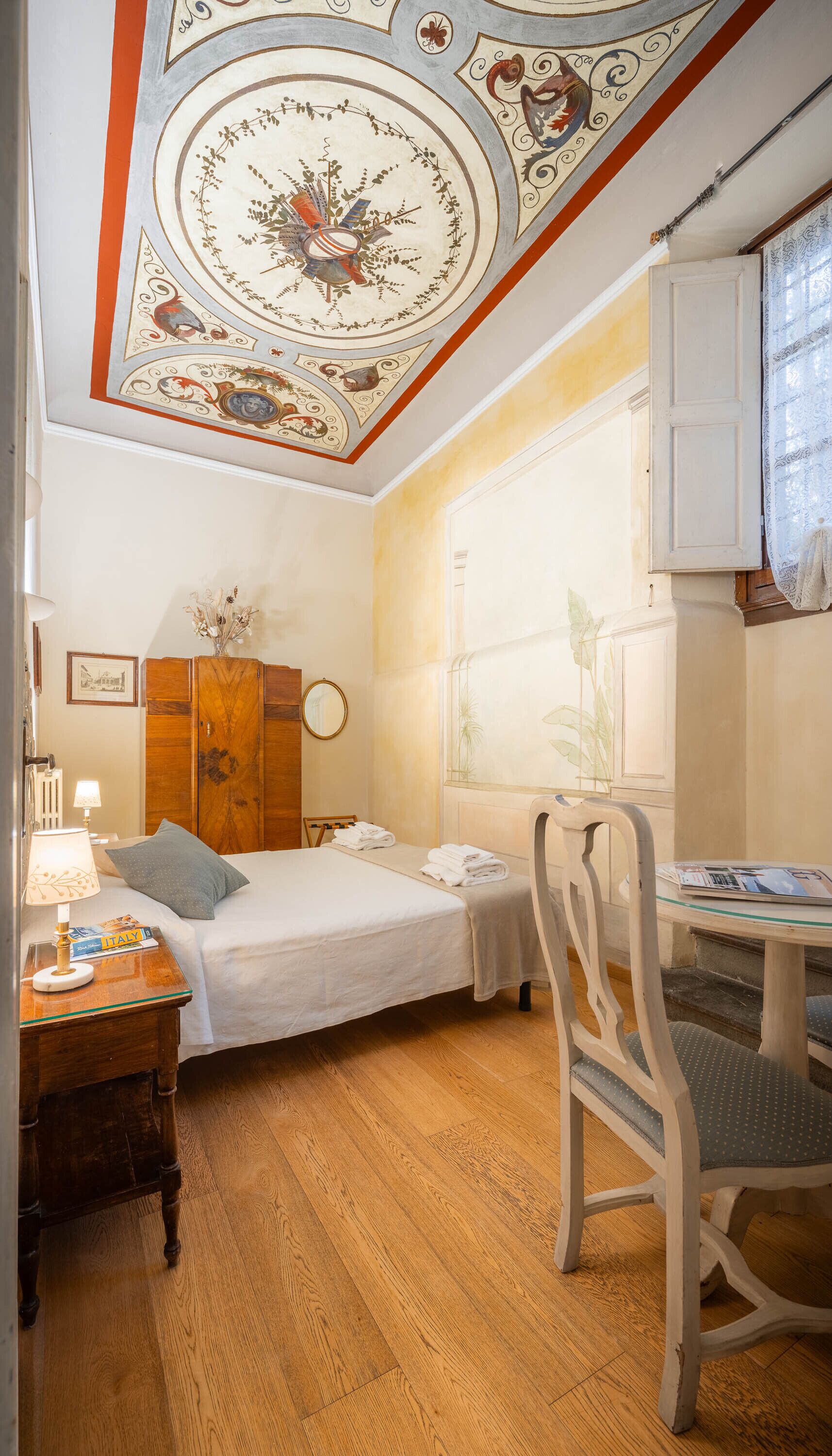 A bedroom with a wooden wardrobe, a bed with a white duvet, a desk with a chair, and a window with a view of plants.