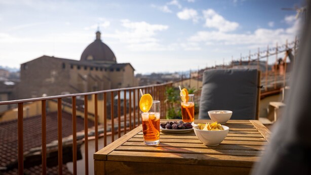 A wooden table with drinks and snacks on a balcony overlooking a cityscape with a prominent dome building. - Novella House (Florence)