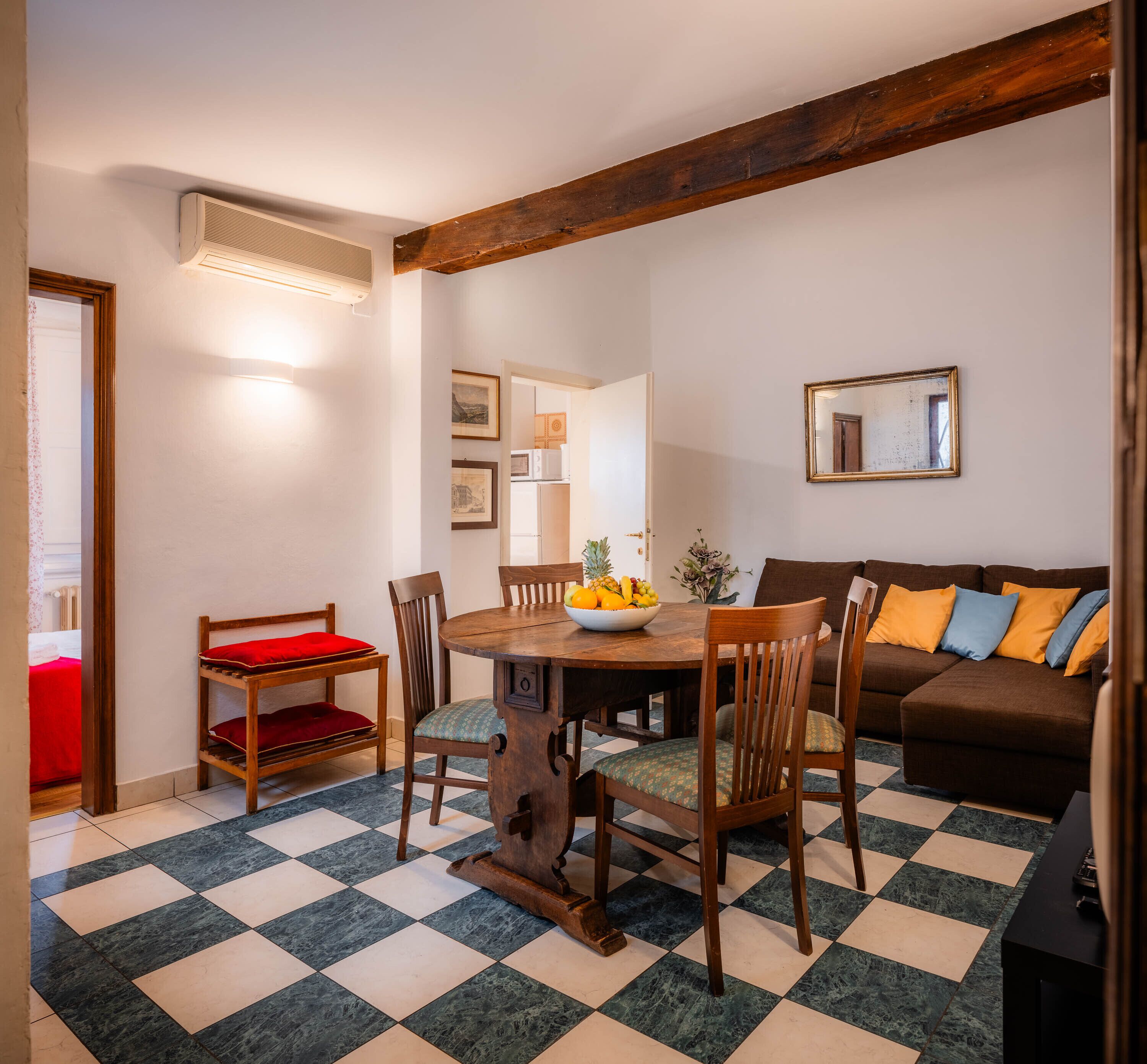 Un coin repas avec une table en bois, des chaises et un saladier rempli de fruits. On aperçoit un canapé marron et un banc rouge à l’arrière-plan.