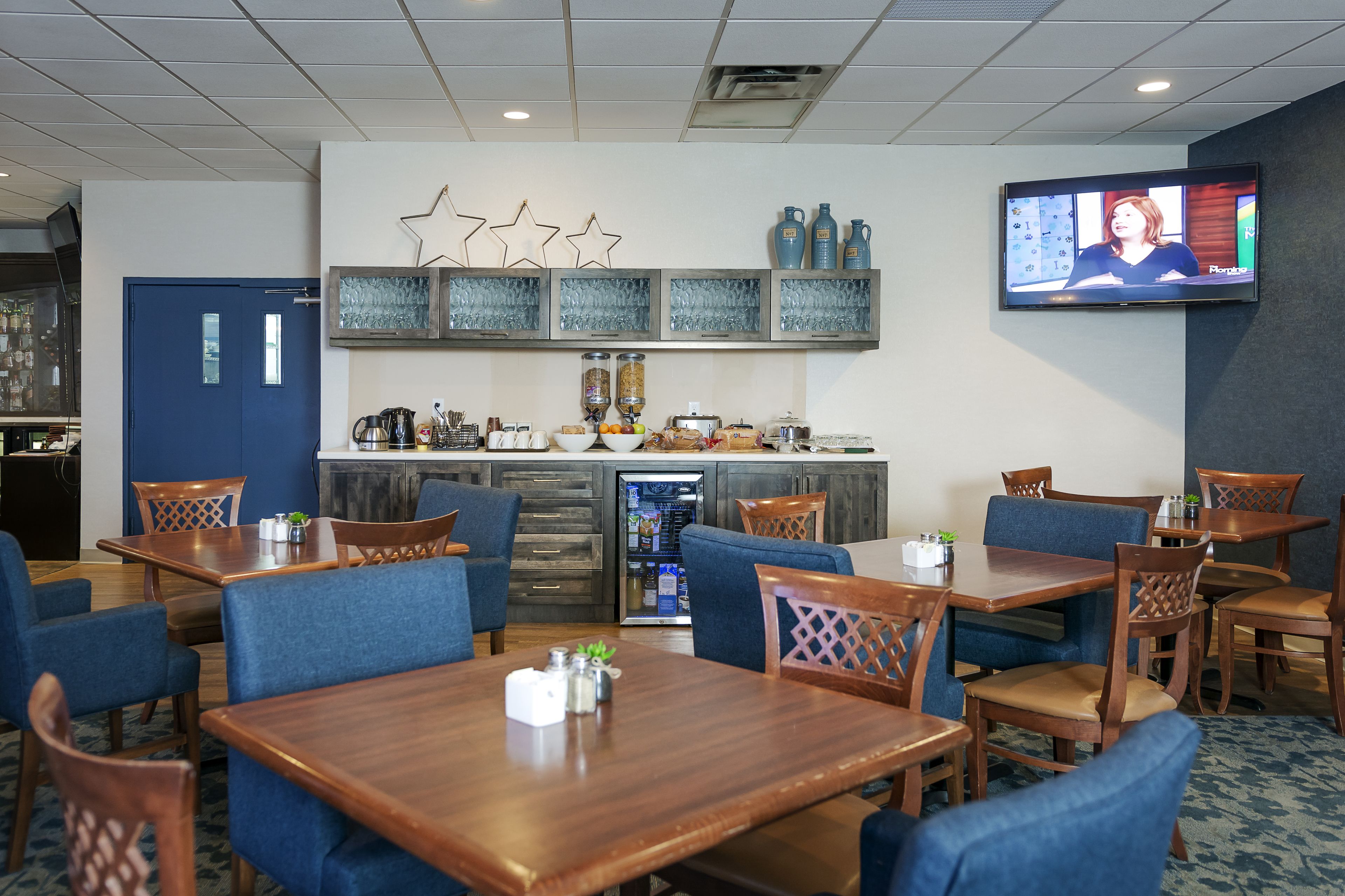 Un restaurant avec des tables en bois et des chaises bleues, un coin cuisine avec un réfrigérateur et un téléviseur fixé au mur.