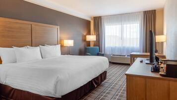 A hotel room with a large bed, two bedside lamps, a desk with a coffee maker, a TV, and an air conditioning unit.