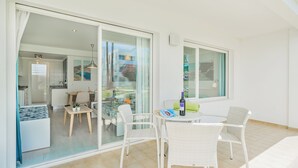 Una habitación moderna y bien iluminada con un vidrio que da a un balcón, muebles de mimbre blanco y una mesa de comedor con una botella de vino.