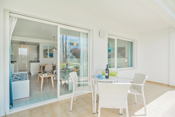 A modern, well-lit room with a glass door leading to a balcony, white wicker furniture, and a dining table with a bottle of wine. - Apartamentos Habitat (Pollenca)