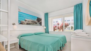 Una habitación con dos camas, un escritorio, un ventanal con cortinas, un cuadro de una escena costera y una zona de baño.
