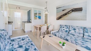 Una sala moderna con sofá, una mesa de comedor con sillas, una zona de cocina y un amplio ventanal con vista al mar.