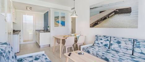 A modern living room with a sofa, a dining table with chairs, a kitchen area, and a large window with a view of the sea.