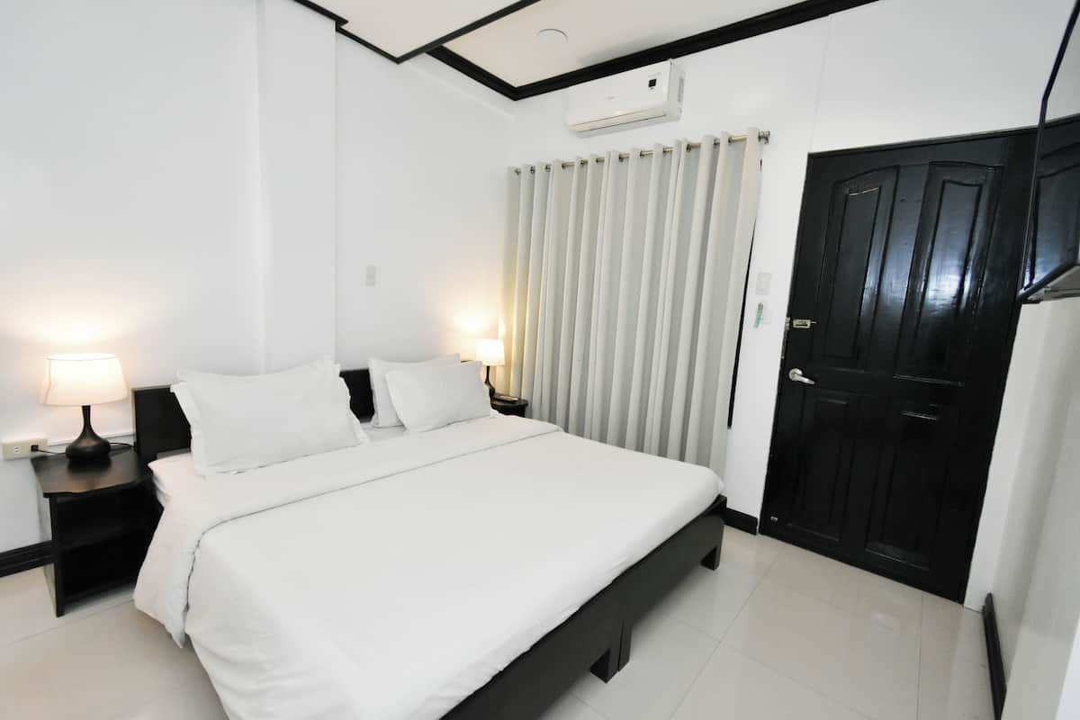 Quarto de hotel com uma cama grande, duas mesas de cabeceira com abajures, uma porta preta e uma janela com venezianas verticais.