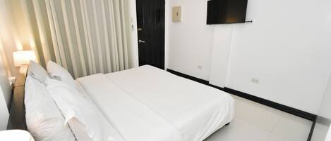 A hotel room with a large bed, a bedside table, a lamp, a door, and a flat-screen TV mounted on the wall.