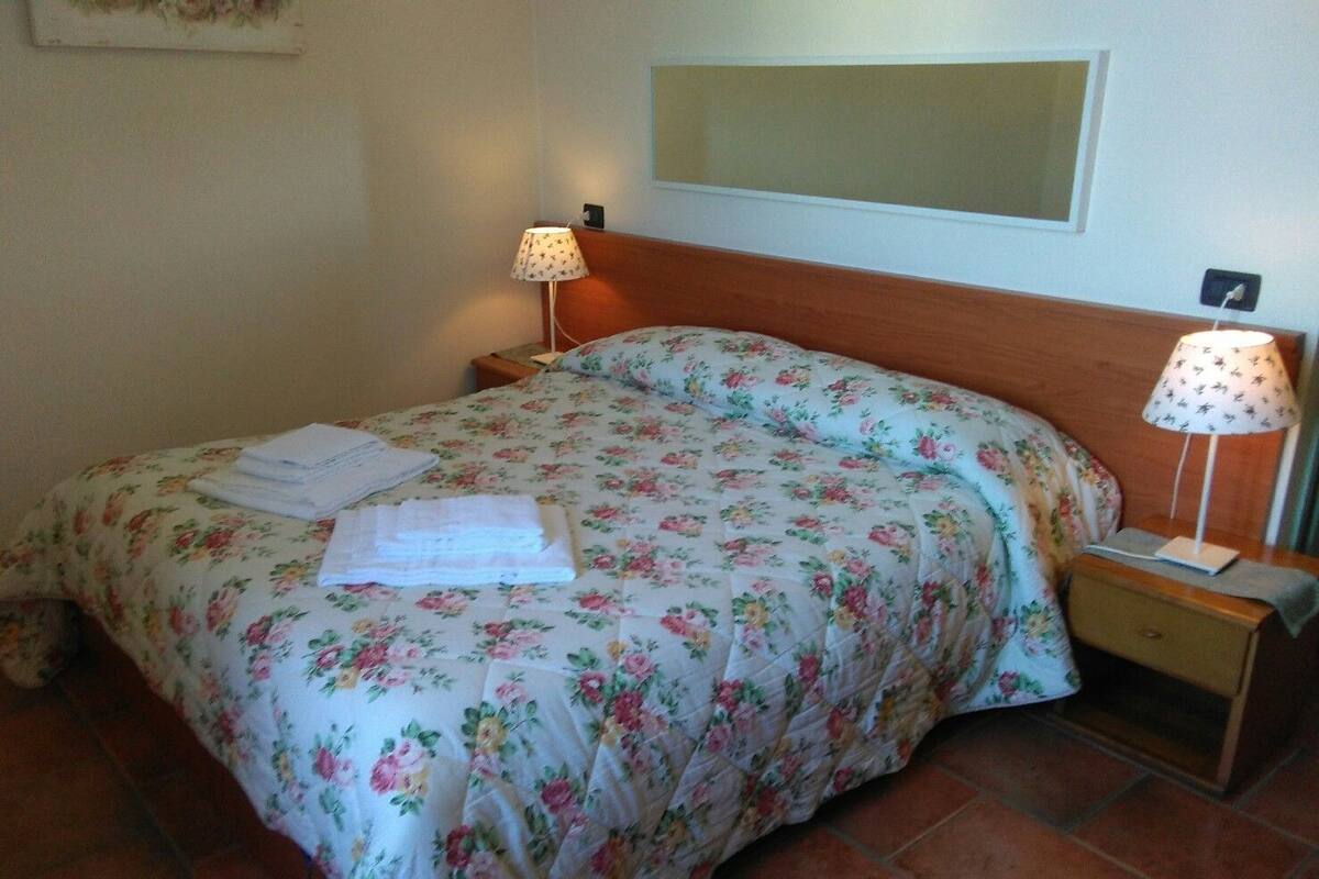 Una cama bien tendida con ropa de cama floral, dos lámparas de noche y una mesita de noche con lámpara.