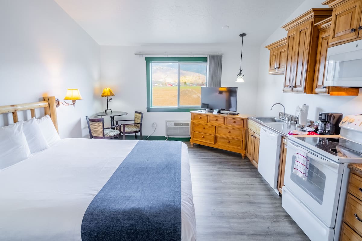 A hotel room with a bed, bedside lamps, a TV, a small table with a chair, a window with a view, a kitchenette with a stove, and wooden cabinets.
