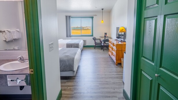 A hotel room with a bed, a desk, a chair, a lamp, a window with a view, and a green door. - Teton Valley Motel (Victor)