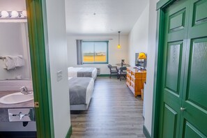 A hotel room with a bed, a desk, a chair, a lamp, a window with a view, and a green door. - Teton Valley Motel (Victor)