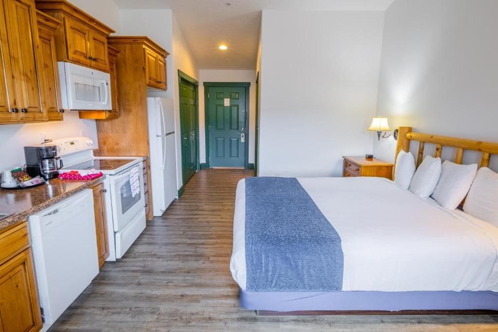 A hotel room with a bed, a bedside table with a lamp, a kitchen area with wooden cabinets, a microwave, and a refrigerator.