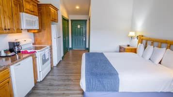 A hotel room with a bed, a bedside table with a lamp, a kitchen area with wooden cabinets, a microwave, and a refrigerator.