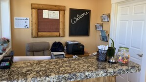 A kitchen counter with a coffee machine, a small seating area, a welcome sign, and a bulletin board. - Teton Valley Motel (Victor)