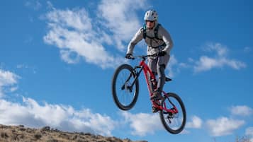 En person på en rød mountainbike i luften, iført hjelm og hvidt tøj, mod en klar blå himmel med spredte skyer.