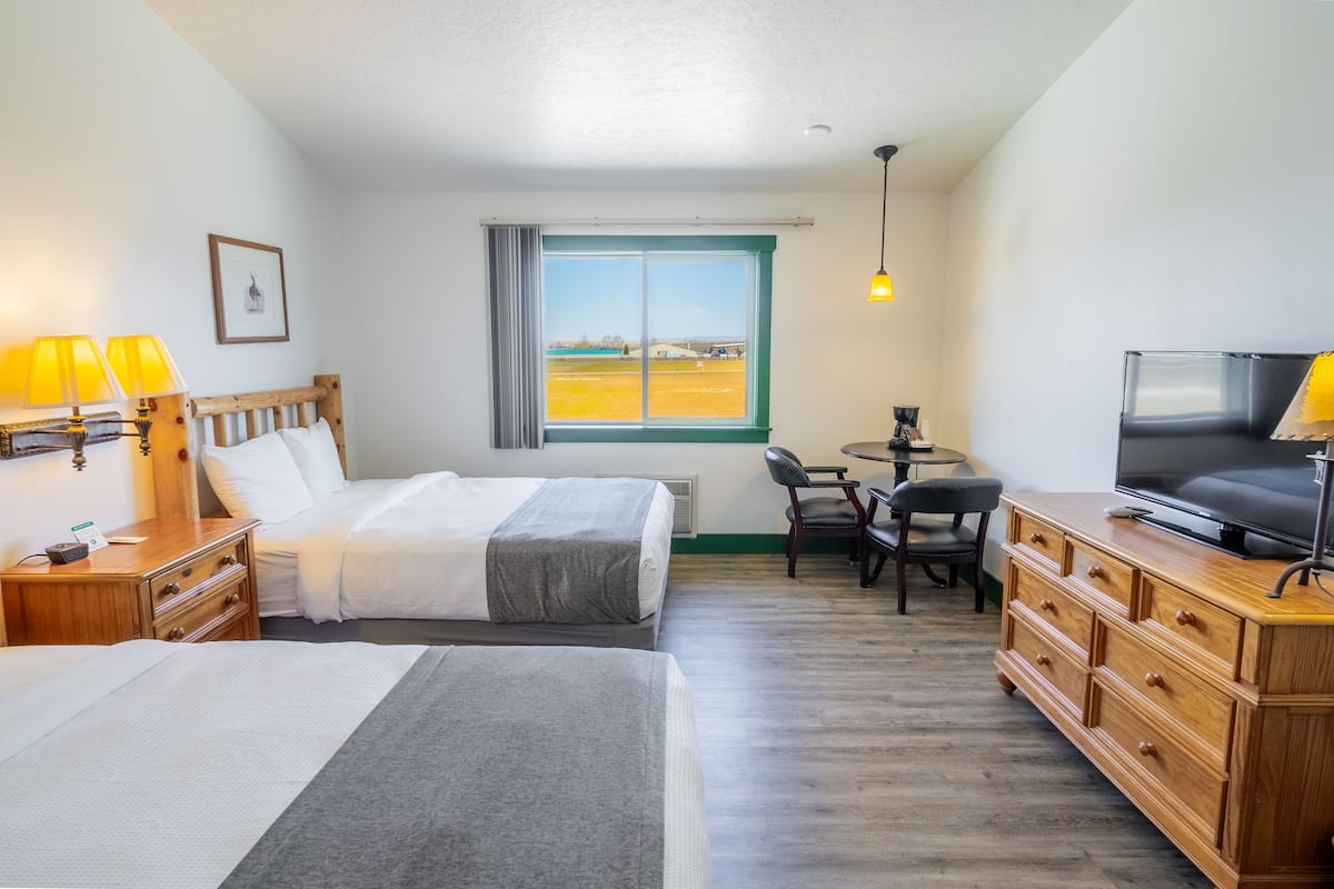 A hotel room with two beds, a desk, a TV, and a view of a field through a window.