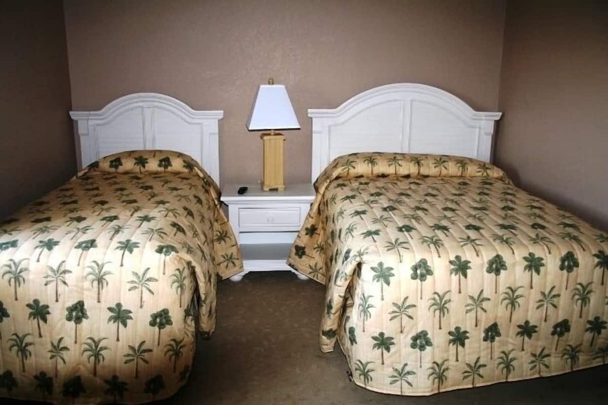 A hotel room with two beds, each with a palm tree patterned bedspread, a white headboard, a bedside table with a lamp, and a small drawer.