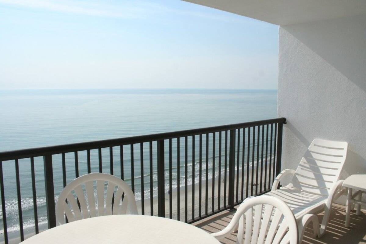 A balcony with white chairs and a table, overlooking a beach and the ocean.