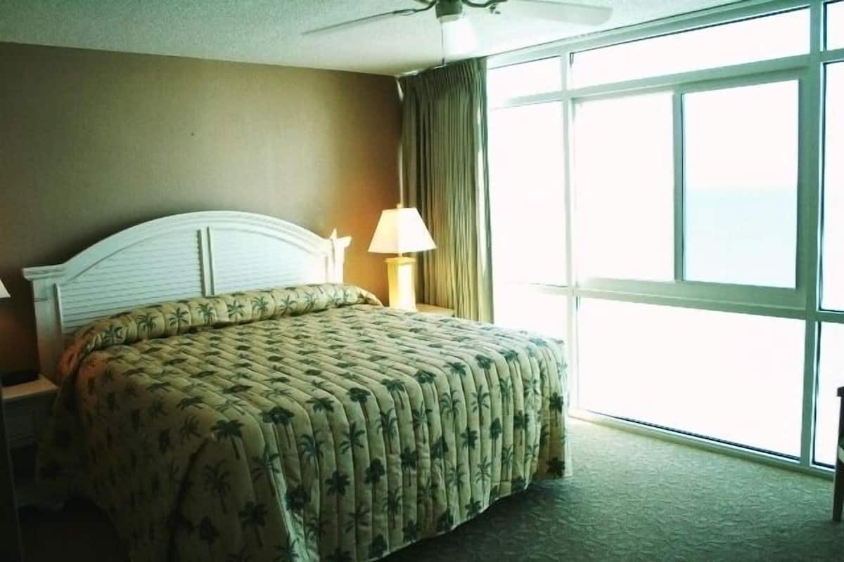 A hotel room with a large bed, a ceiling fan, a nightstand, a lamp, and a view of the ocean through large windows.