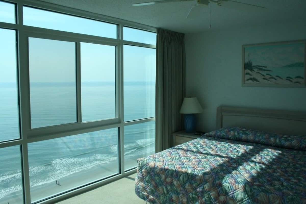 A hotel room with a large window offering a view of the ocean, a bed with a patterned bedspread, a nightstand with a lamp, and a painting on the wall.