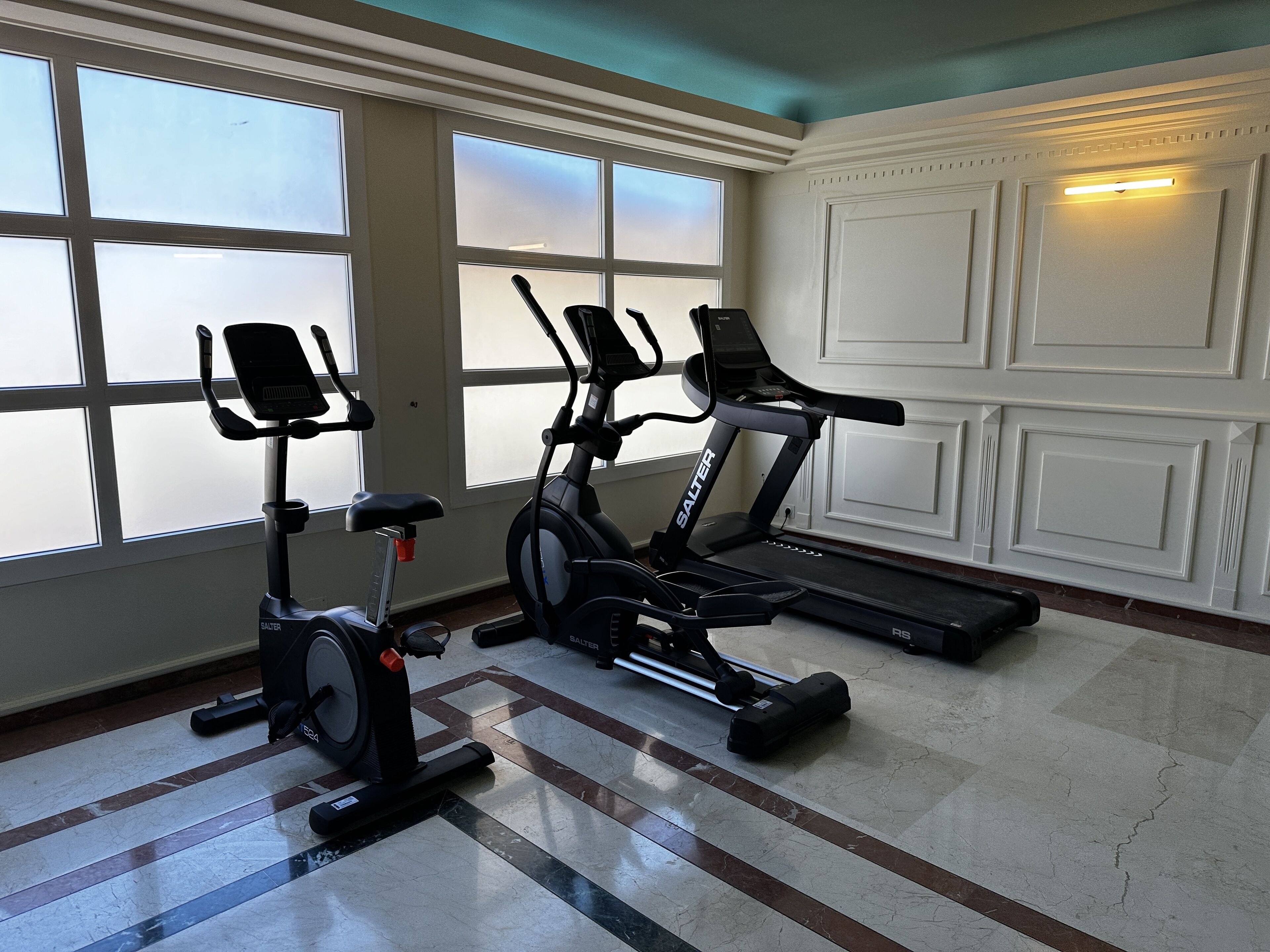 Un gimnasio con dos bicicletas estáticas y una cinta de correr, ventanas amplias y piso de baldosas.