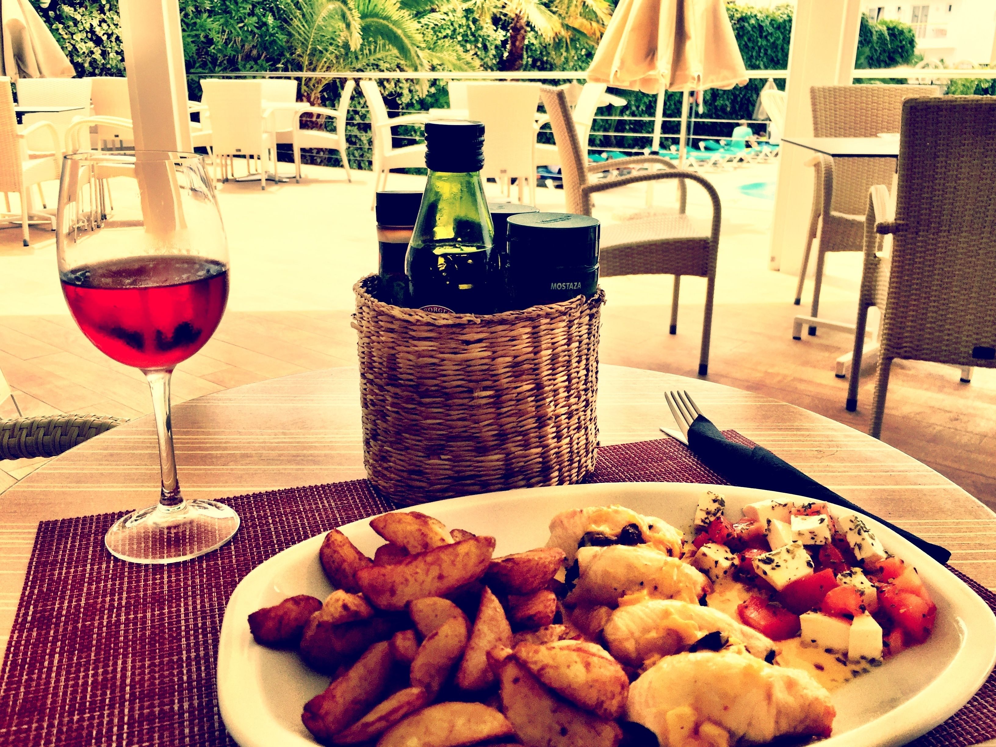 A dining table with a plate of food, a glass of red wine, and a wicker basket with bottles.