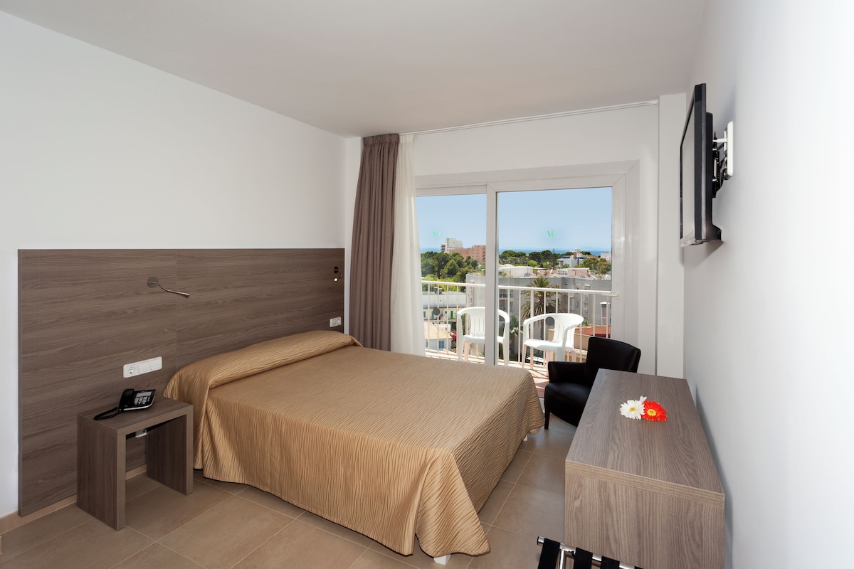 Habitación de hotel con cama, escritorio, silla, televisión y balcón con vistas.