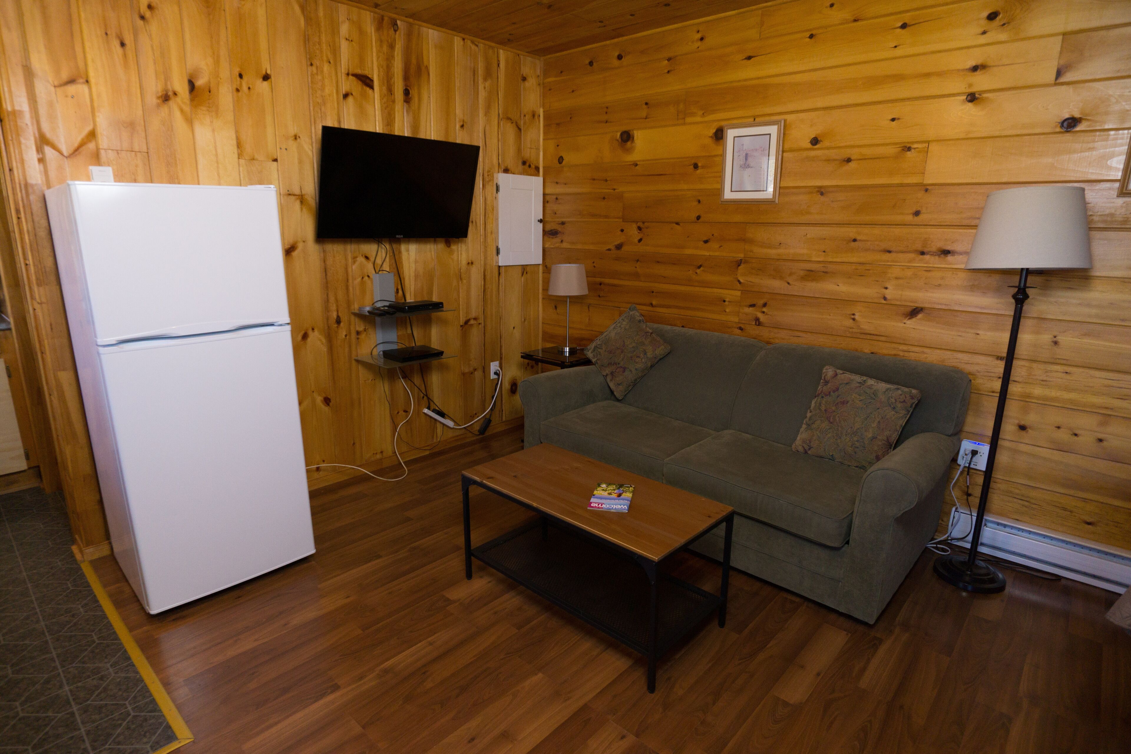 Una habitación con paneles de madera, un refrigerador blanco, un televisor montado en la pared, un sofá con un cojín, una mesa de centro y una lámpara de pie.