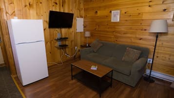 A wooden-paneled room with a white refrigerator, a television mounted on the wall, a sofa with a cushion, a coffee table, and a floor lamp.