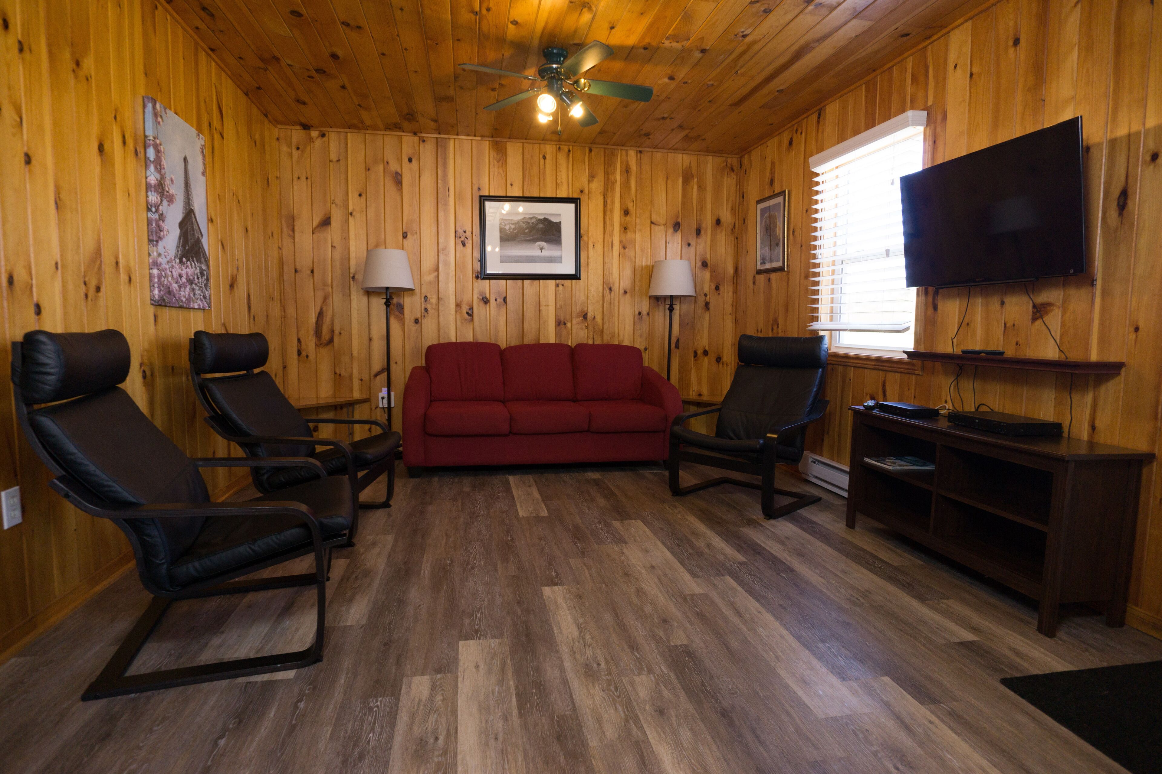 A wooden-paneled room with a red sofa, two black chairs, a television, and two floor lamps.