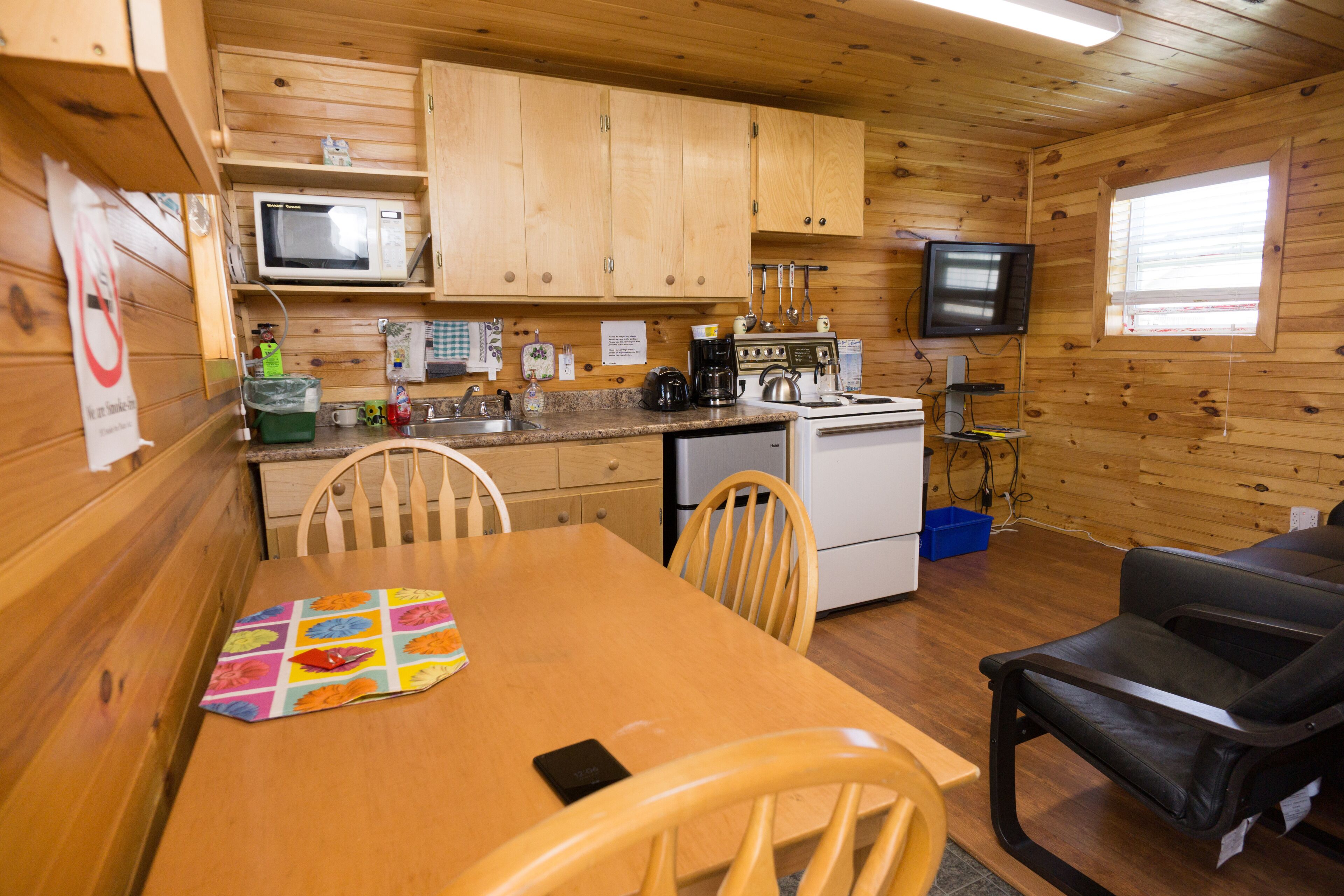 A wooden cabin with a kitchen area including a microwave, stove, and sink. There is a dining table with chairs and a television mounted on the wall.