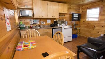 A wooden cabin with a kitchen area including a microwave, stove, and sink. There is a dining table with chairs and a television mounted on the wall.