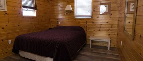 A wooden cabin bedroom with a bed, a nightstand, a lamp, and two windows.
