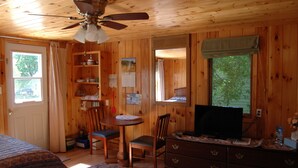 Una habitación de cabaña de madera con una cama, una mesa de comedor con sillas, un televisor y un ventilador de techo.