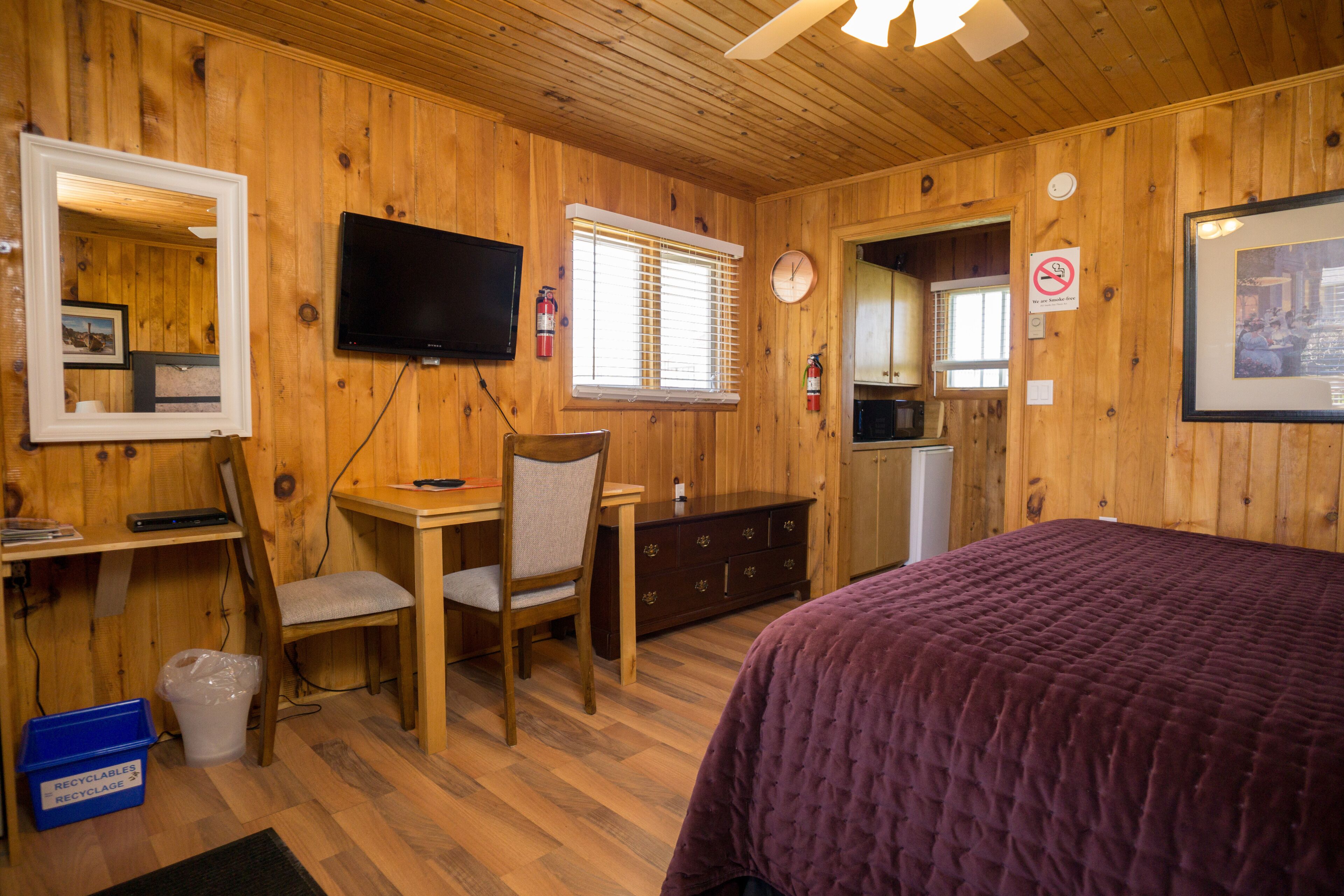 Una habitación de cabaña de madera con cama, escritorio, silla, televisor y una pequeña cocina.