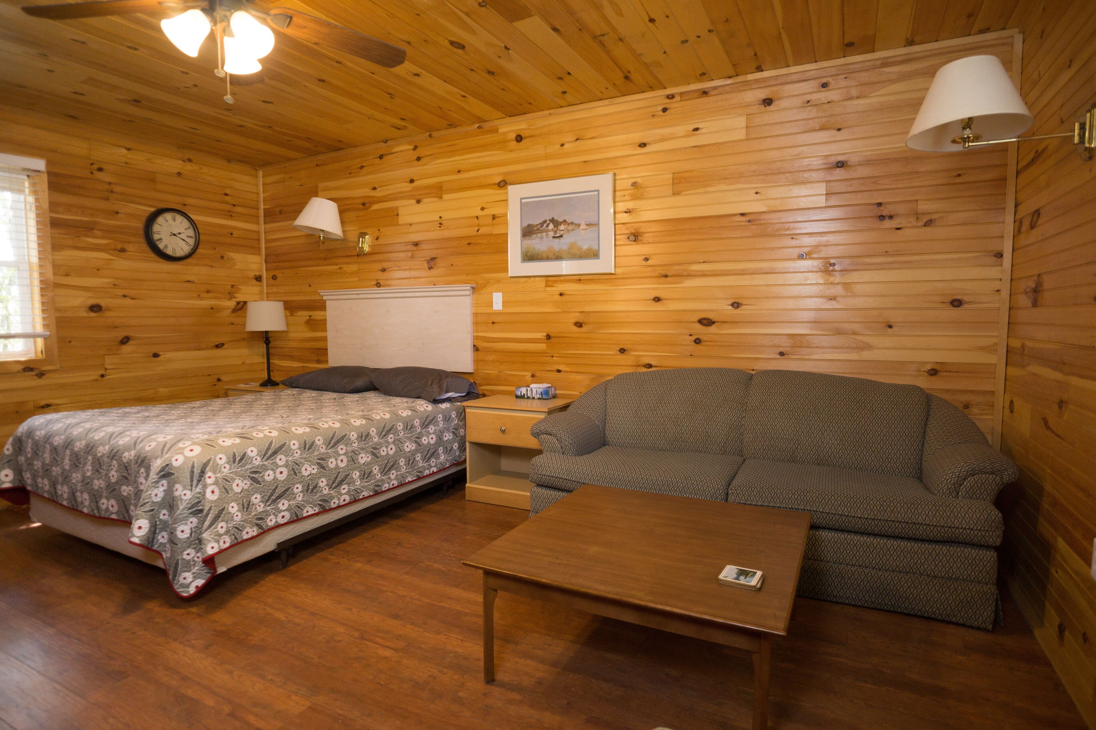 A wooden cabin room with a bed, a sofa, a table, a lamp, and a clock on the wall.