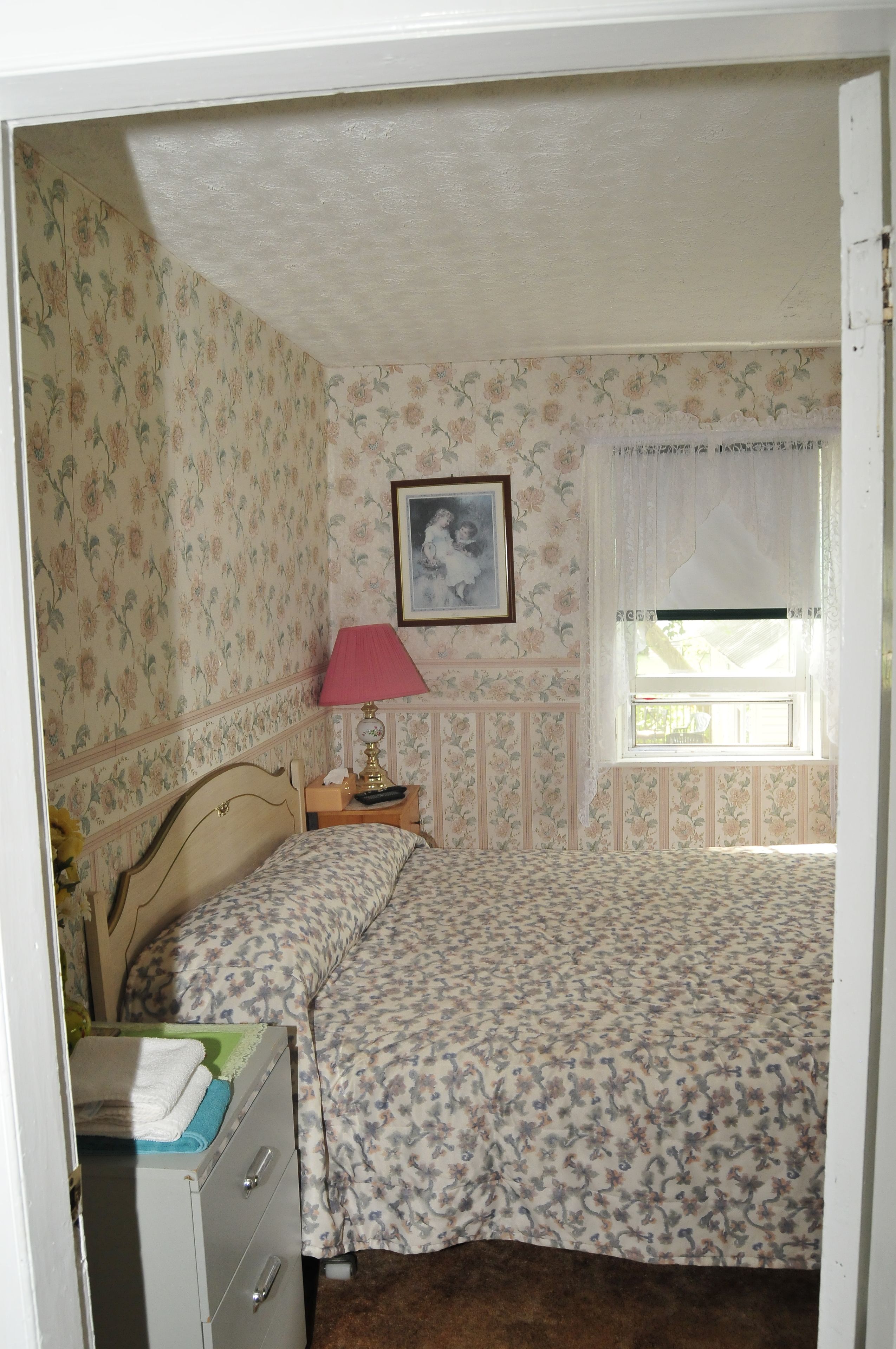 A bedroom with floral wallpaper, a bed with a floral bedspread, a nightstand with a lamp, and a window with curtains.