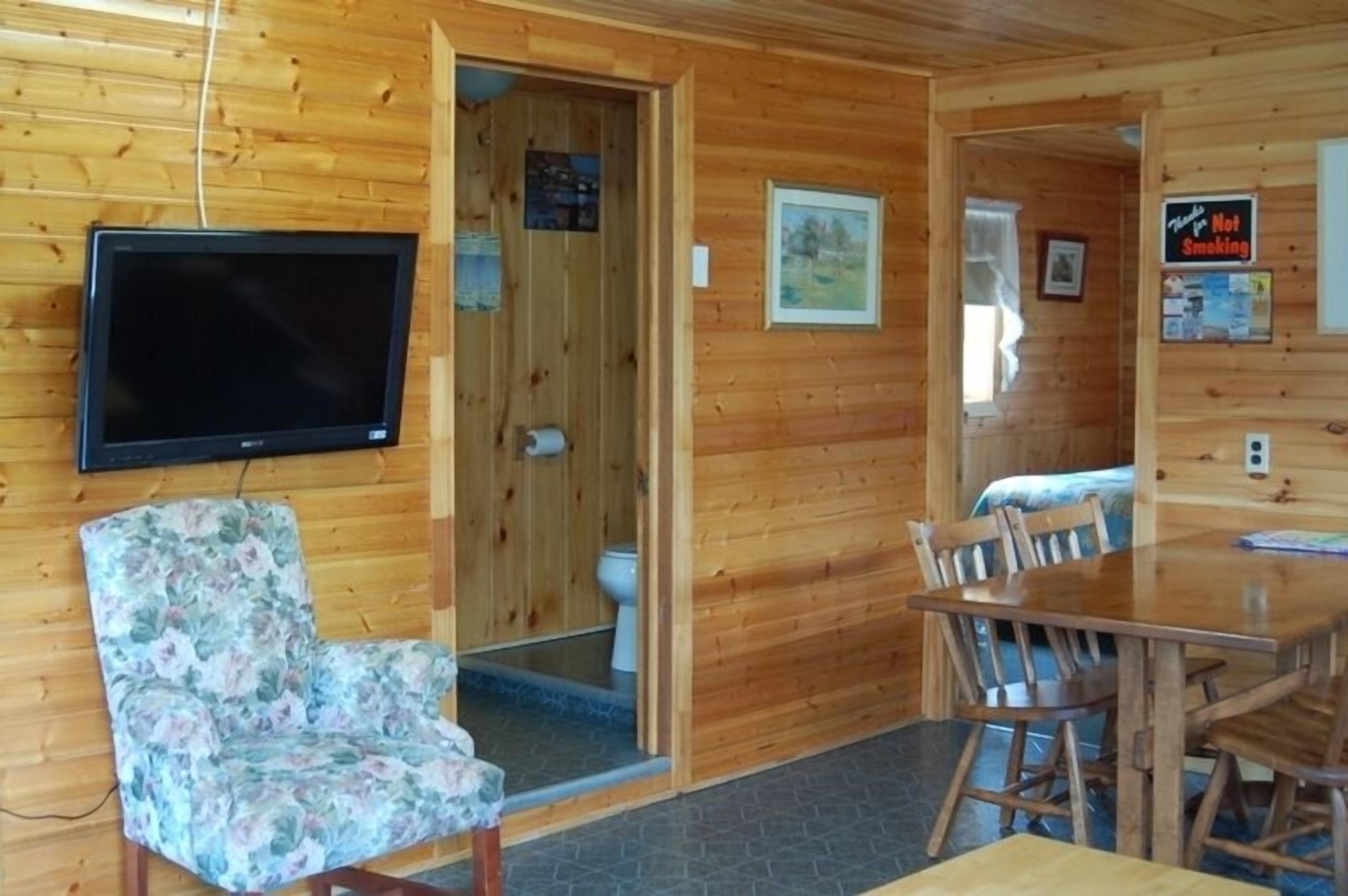 A wooden cabin interior with a television, a floral chair, a wooden dining table, and a small bathroom with a toilet and sink.