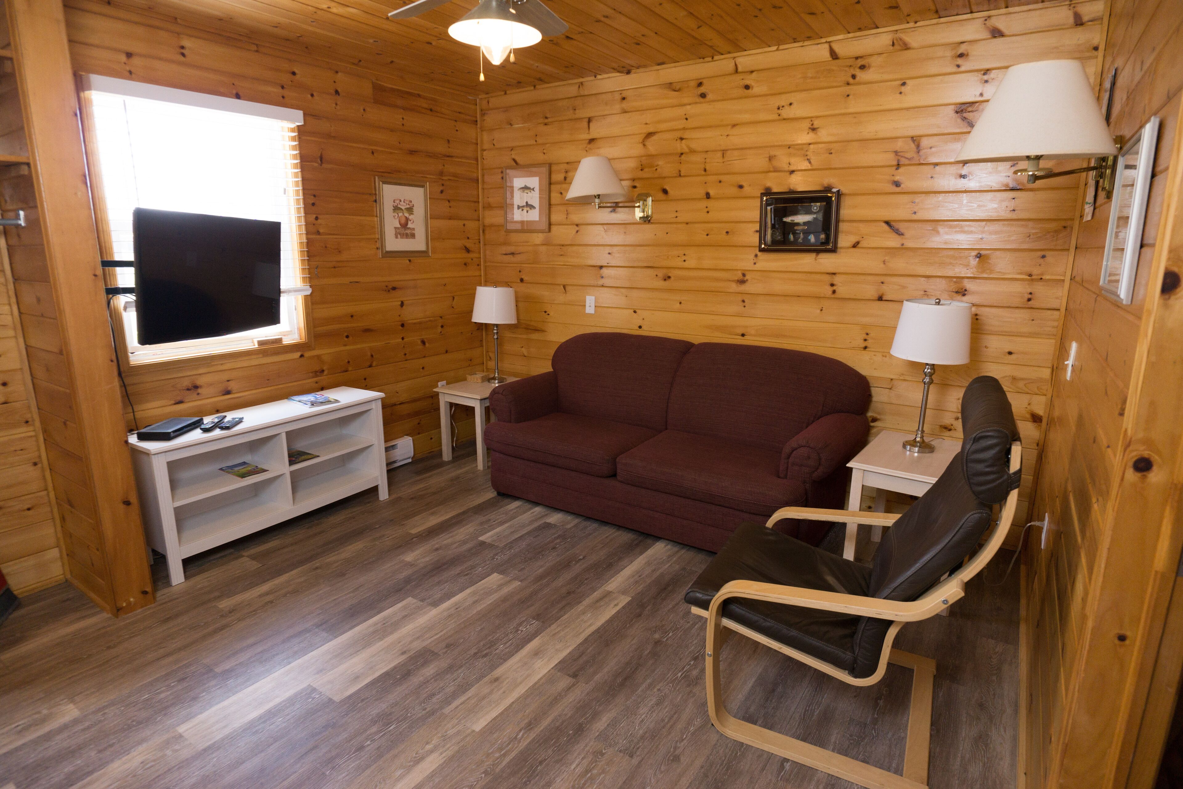 A cozy cabin interior with a wooden floor, a red sofa, a black chair, a small white table, and a white shelf with a TV and remote control.