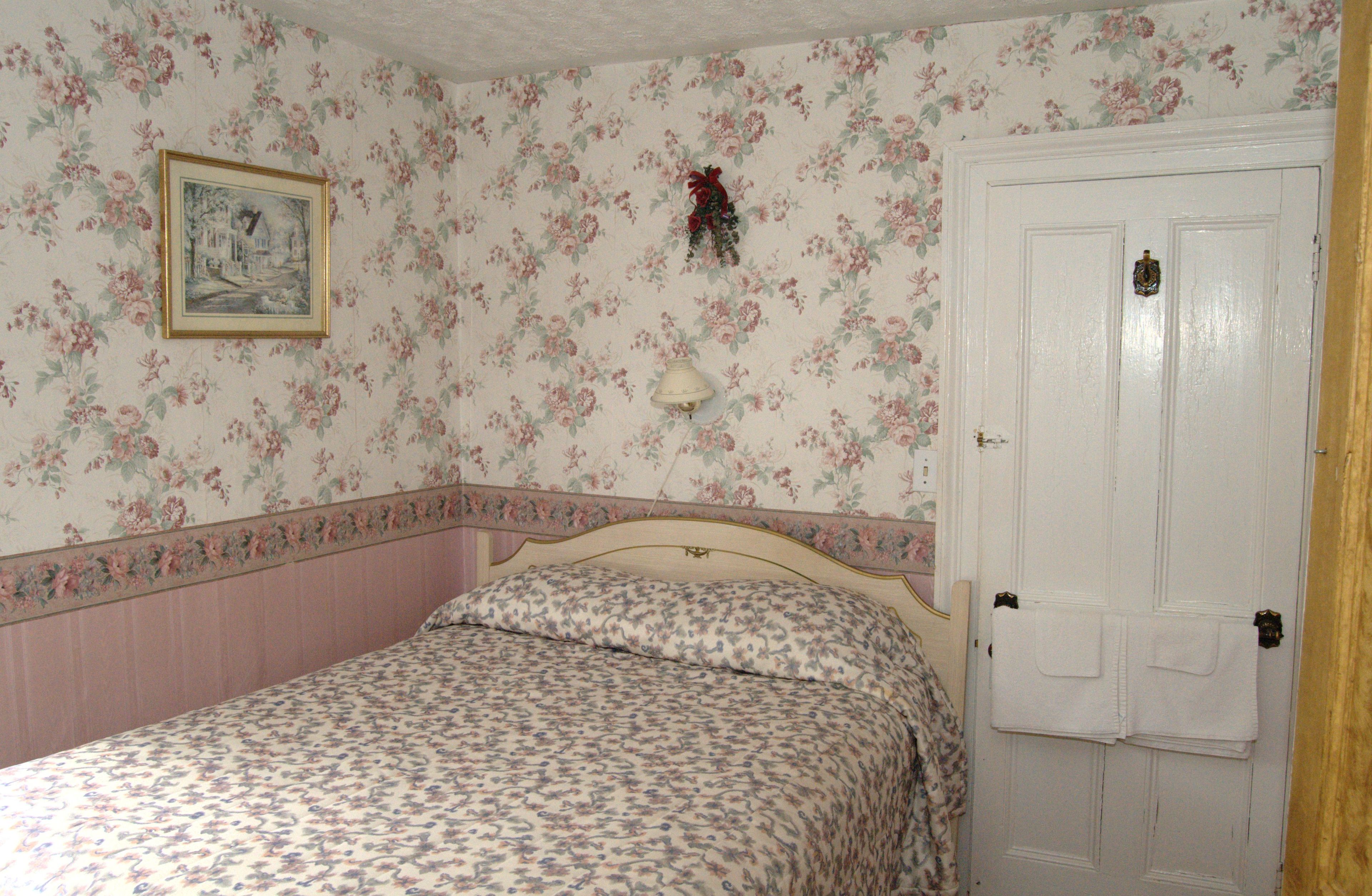 A bedroom with floral wallpaper, a bed with a floral bedspread, a nightstand, a wall-mounted lamp, and a door with a towel rack.