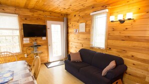 Interior acogedor de una cabaña con un sofá azul oscuro, paredes de madera, un televisor y un comedor con mesa y sillas.