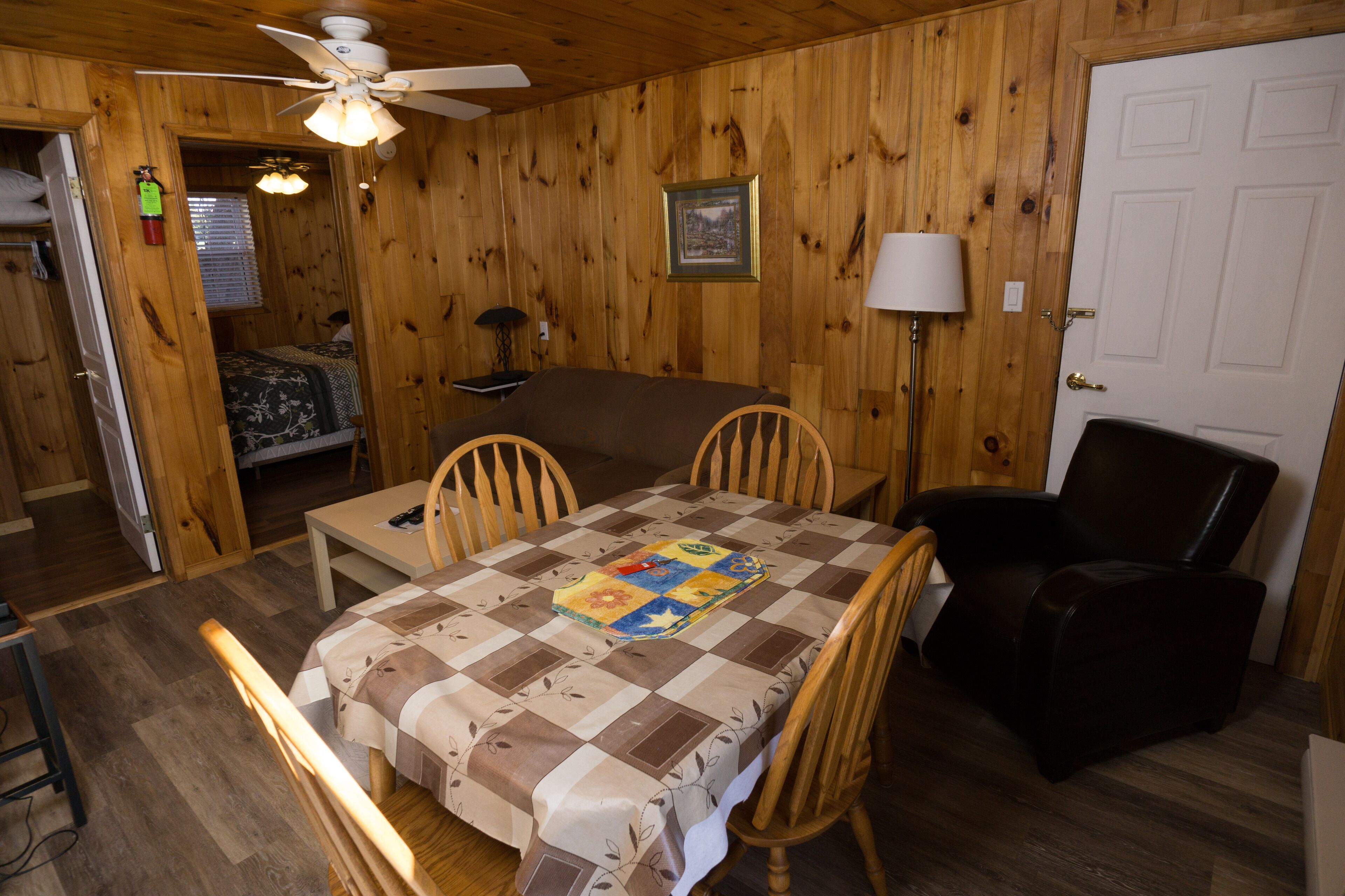 Una habitación acogedora con detalles de madera, una mesa de comedor con sillas, un sillón de cuero negro, un ventilador de techo, un cuadro en la pared y una puerta.