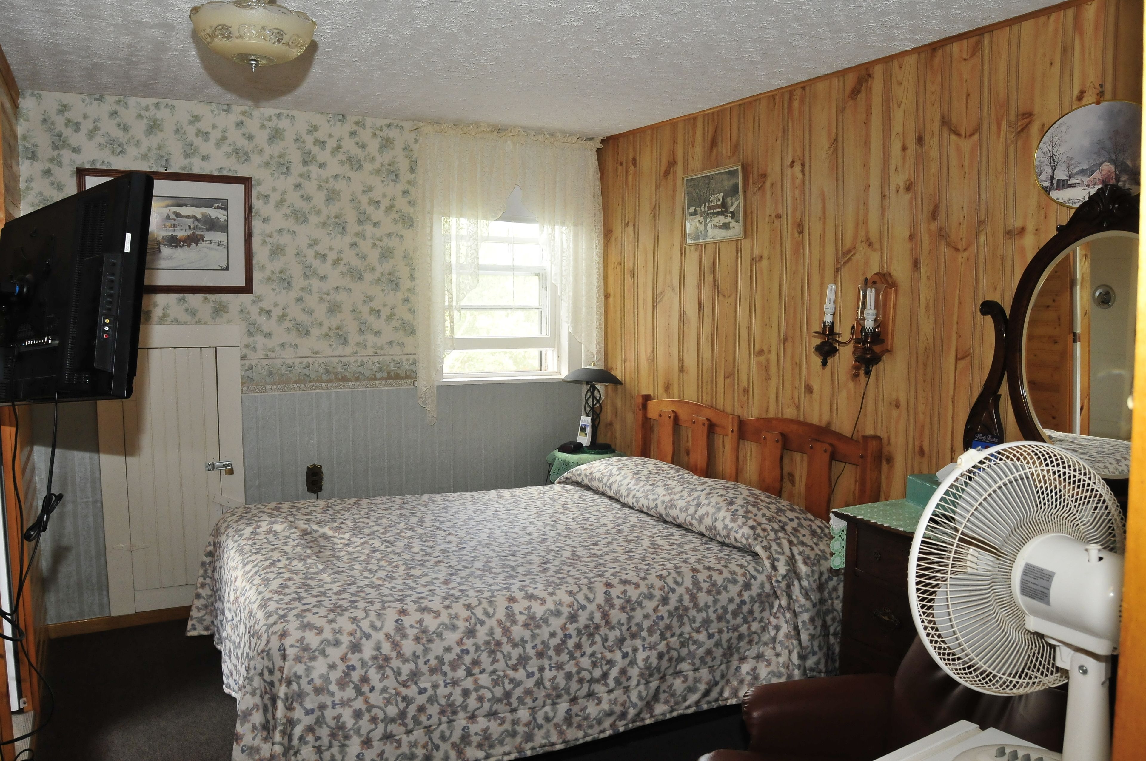 Een slaapkamer met een houten hoofdbord, een bed met bloemenbeddengoed, een nachtkastje met een lamp, een aan de muur bevestigde televisie en een staande ventilator.