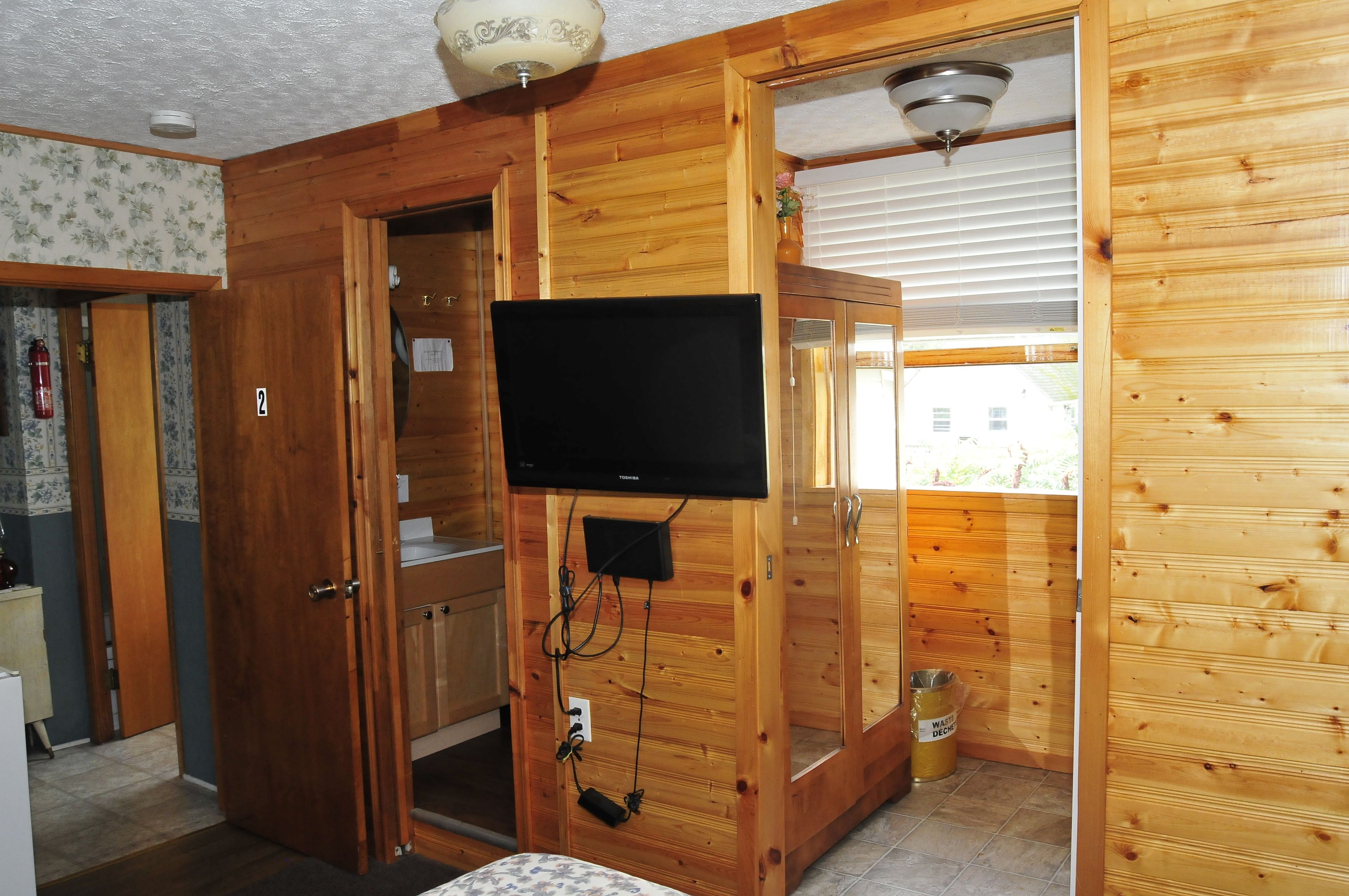 A wooden cabin with a television mounted on a wall, a door leading to a bathroom, and a window with blinds.