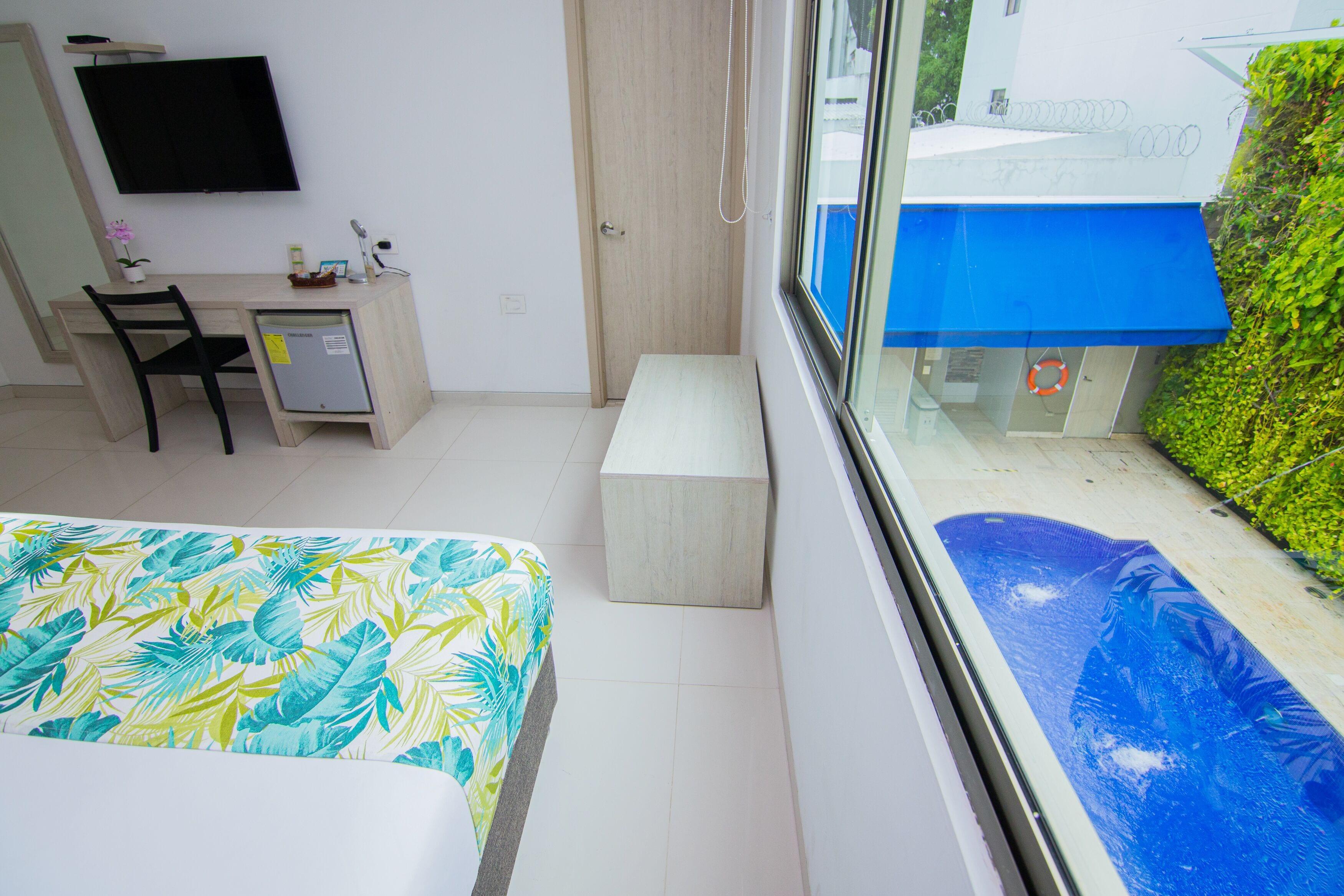 Un dormitorio con cama, escritorio, silla, televisor y ventana con vista a una piscina y vegetación.