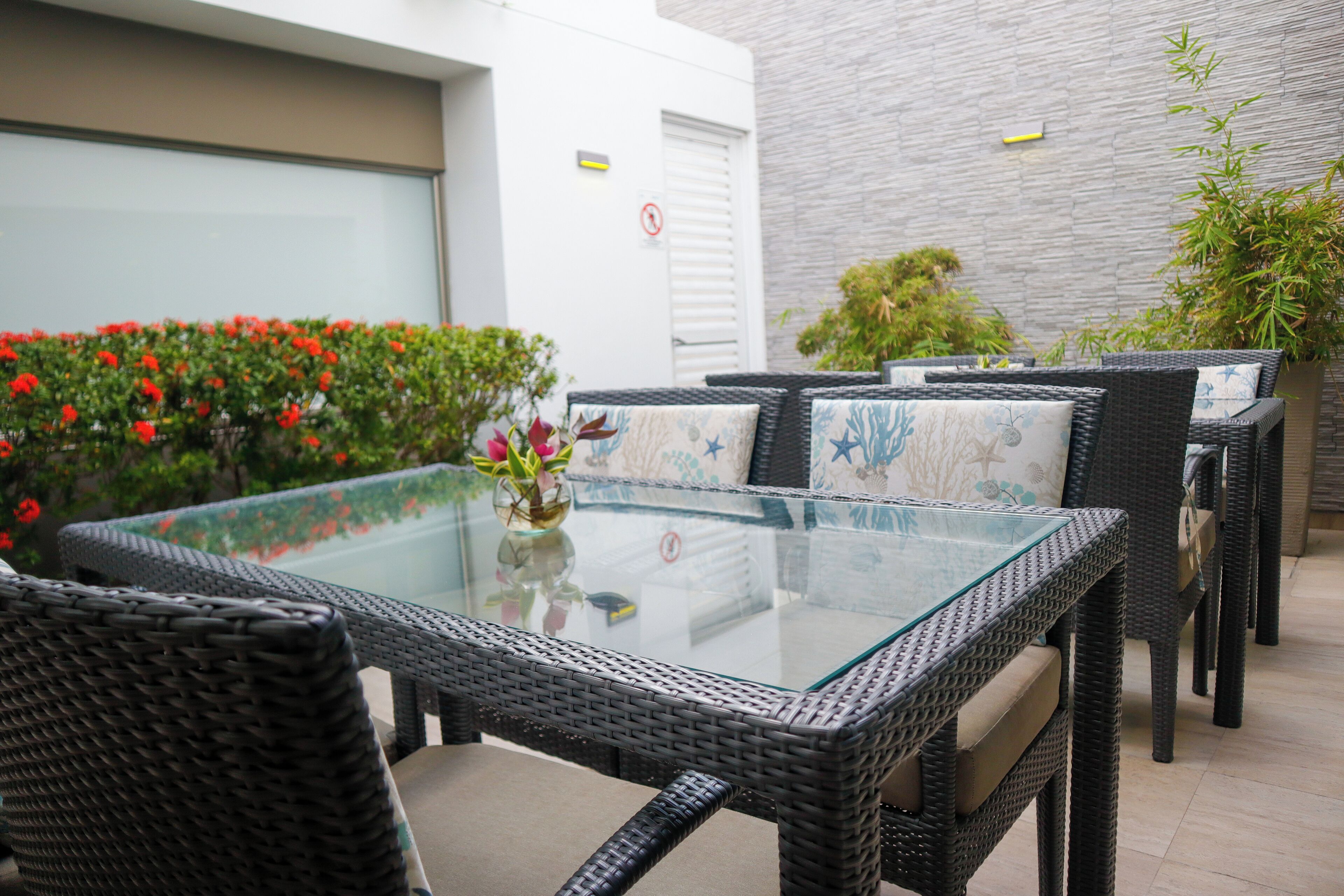 An outdoor seating area with wicker furniture and a glass-top table, surrounded by plants and a building wall.