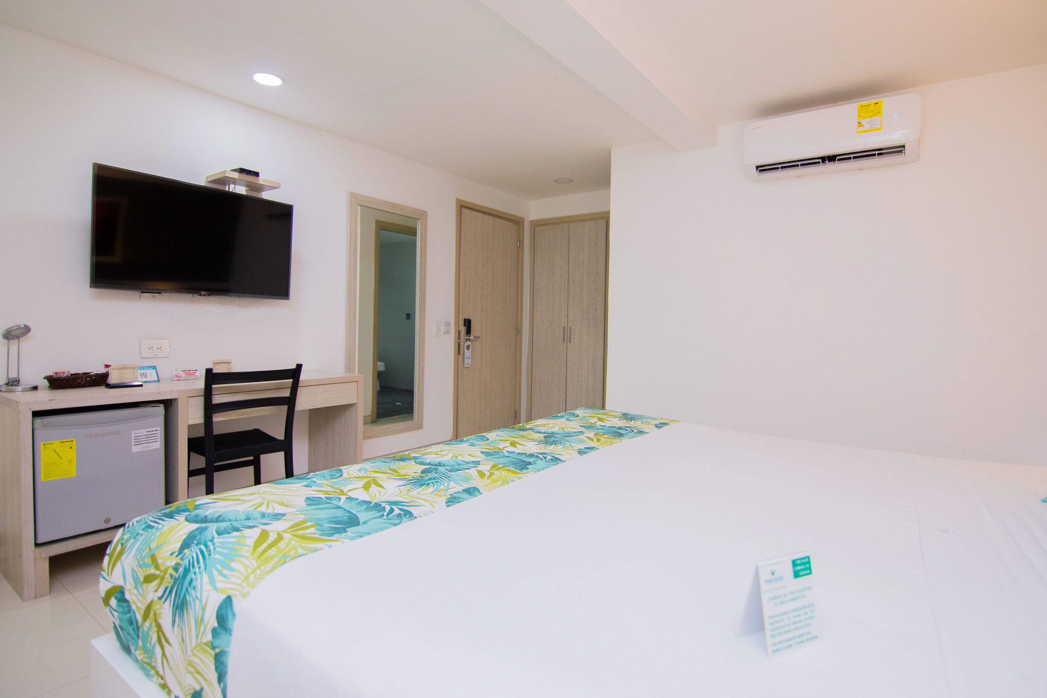 A hotel room with a bed, a desk with a computer, a TV, and an air conditioning unit.